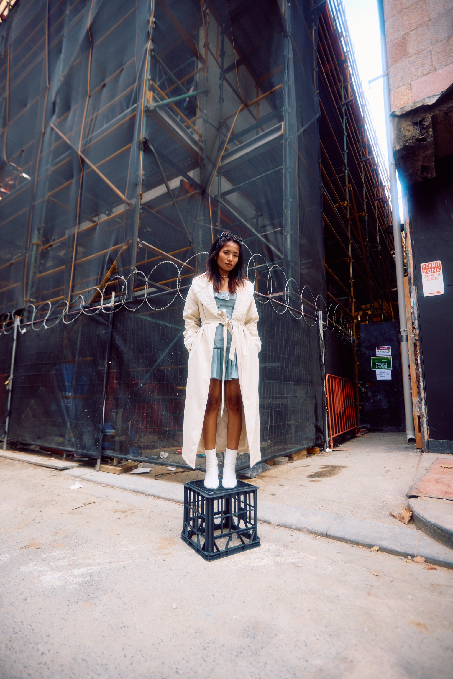 A young woman stands on a black plastic crate in an urban alleyway, wearing a long trench coat and white boots, posing for a fashion photograph.
